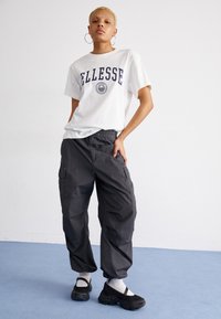 Camiseta blanca de manga corta con logo azul de "Ellesse", pantalones cargo gris oscuro con bolsillos y zapatillas deportivas negras con calcetines blancos.