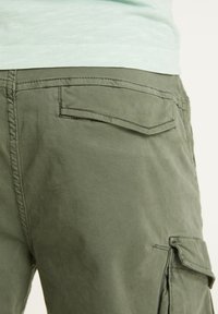 Gros plan sur la poche et le rabat d’un pantalon cargo vert olive avec des détails de couture, porté avec une chemise de couleur claire.