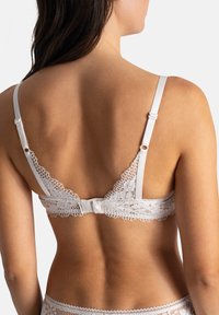 Soutien-gorge en dentelle blanche avec des bretelles réglables, des bords en feston et une fermeture crochet-oeil au dos, orné de motifs floraux délicats.