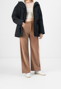 Veste matelassée noire avec capuche, doublure crème et poches zippées ; associée à un pantalon large en velours côtelé côtelé marron et des baskets blanches.