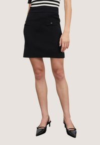 Musta mini-hame, jossa on kapea vyötärö, kaksi etutaskua ja kultaiset yksityiskohdat. Yhdistetty mustiin kärkitossuihin. Näyttävä kangas.