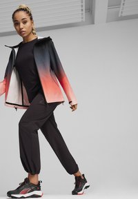 Giacca con gradiente nero e arancione, top nero e pantaloni cropped neri; materiale leggero con polsini elasticizzati e sneaker sportive con dettagli rossi.