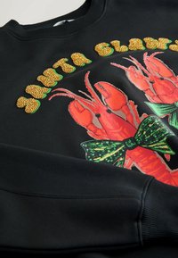 Czarna bluza z grafiką czerwonego homara, z zielonymi kokardkami i złotymi koralikami, podkreślająca teksturowany wzór w jaskrawych kolorach.
