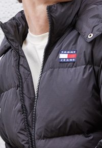 Czarna kurtka puchowa z zapięciem na zamek i wysokim kołnierzem. Posiada tkaninową naszywkę z logo w czerwonych i białych akcentach. Cała kurtka ma pikowaną fakturę.