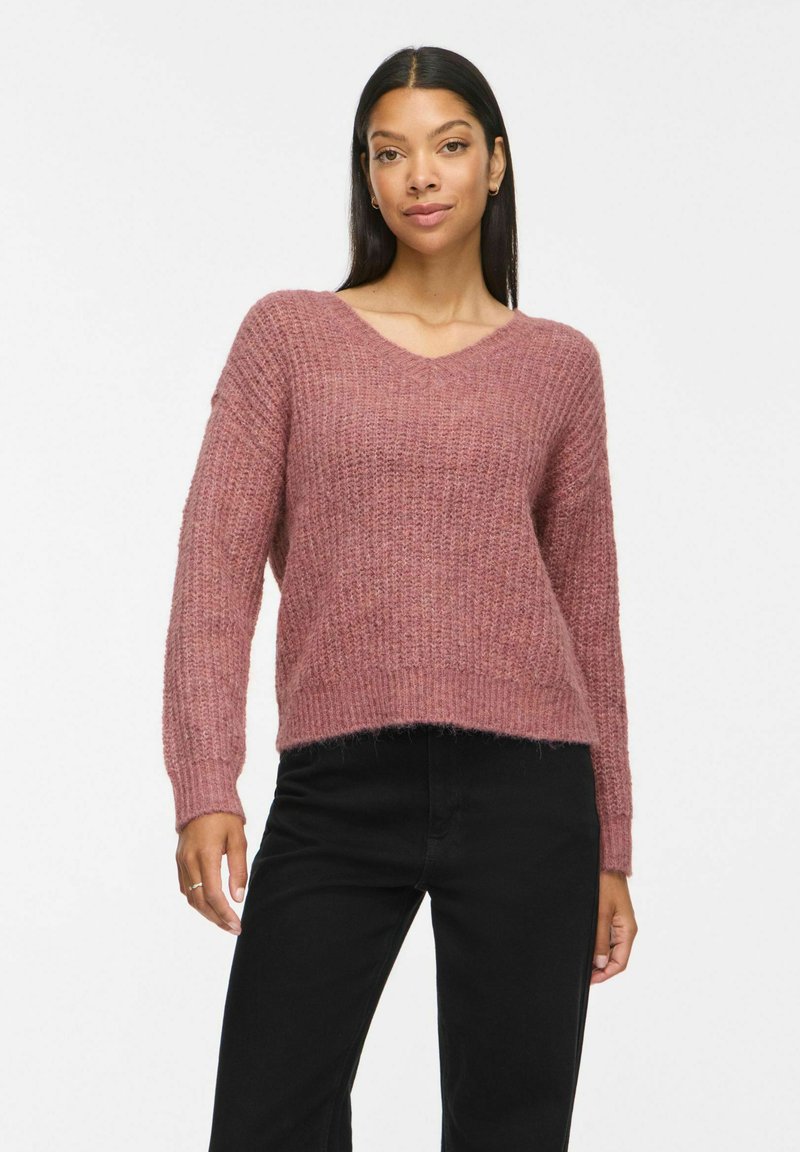 Femme aux longs cheveux noirs portant un pull en maille rose à col en V et un pantalon noir, debout devant un fond clair uni.