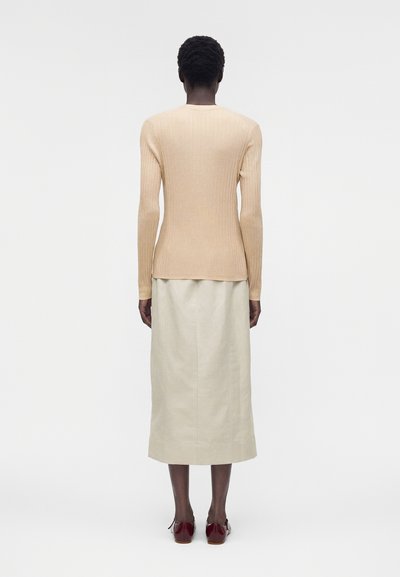Άτομο με κοντά μαλλιά που φοράει μπεζ ριπλό πουλόβερ, κρεμ μίντι φούστα και μπορντό παπούτσια, στέκεται με την πλάτη προς τα πίσω σε απλό φόντο.
