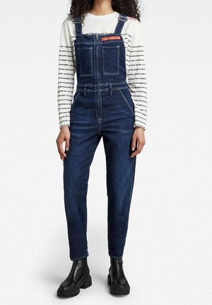 Marineblå denim overalls med en frontlynlås, justerbare stropper og to brystlommer. Sammen med en hvid-langærmet top og sorte støvler.