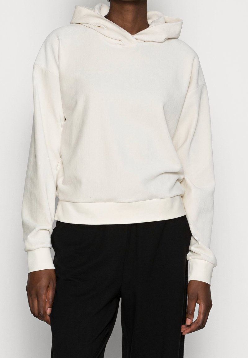 Personne portant un sweat à capuche uni blanc cassé à manches longues et un pantalon noir, montré du cou jusqu'aux hanches.