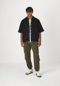 Svart kortärmad skjorta med vit sömnad, oversized fickor; olivgröna cargo-byxor; vita sneakers med svarta ränder; mörka solglasögon.