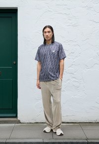 Mann mit langen Haaren, der ein gemustertes lila T-Shirt, beige Cordhosen und weiße Turnschuhe trägt, steht vor einer weißen strukturierten Wand neben einer grünen Tür.