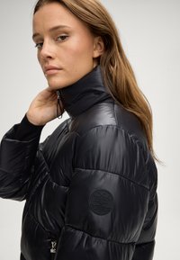 Giacca a vento nera con colletto alto, chiusura con zip e finitura texturizzata. Presenta un patch circolare con il logo sul braccio sinistro.
