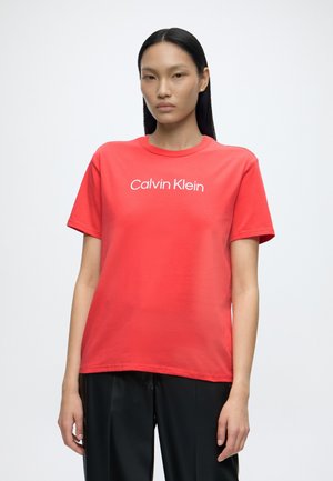 Kobieta o prostych czarnych włosach, ubrana w czerwoną koszulkę Calvin Klein i czarne spodnie, stojąca na jednolitym białym tle.
