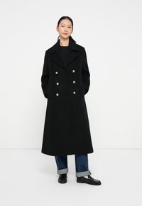 Manteau en laine noir à double boutonnage avec de grands boutons en argent, revers larges et poches latérales ; porté par-dessus un jean retroussé et des chaussures noires.
