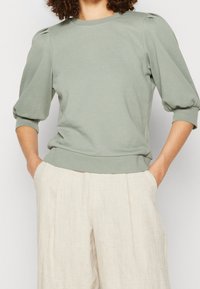 Lysegrønt pullover-top med trekvart puffærmer, rund halsudskæring og ribbet kant, parret med beige vidde bukser.