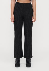 Femme portant un pantalon taille haute noir avec de subtils rayures verticales et des bottines à plateforme noires, se tenant devant un fond uni.