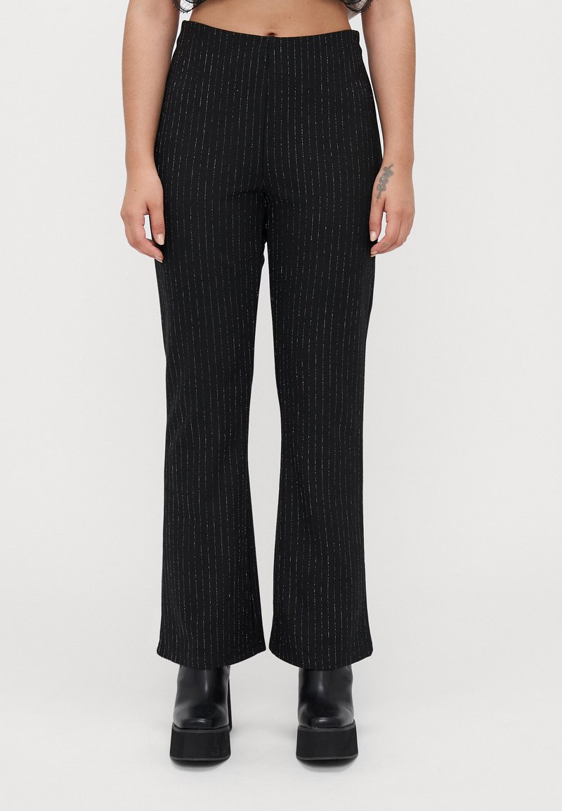 Femme portant un pantalon taille haute noir avec de subtils rayures verticales et des bottines à plateforme noires, se tenant devant un fond uni.