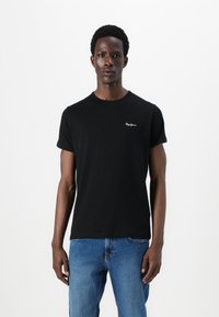 T-shirt nera in cotone con scollatura rotonda e maniche corte, con un piccolo logo bianco sul lato sinistro del petto. Indossata con jeans blu.