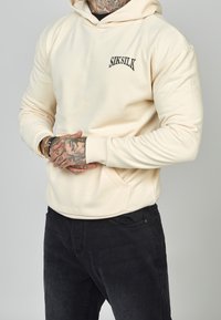 Hombre vistiendo una sudadera crema de SikSilk y jeans negros, con las manos tatuadas entrelazadas a la altura de la cintura, frente a un fondo gris liso.