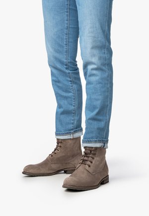 Benen in lichtblauwe opgerolde jeans en bruine suède veterschoenen tot de enkel op een witte achtergrond.