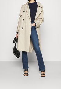 Beige Trenchcoat mit doppelt geknöpftem Front, großen schwarzen Knöpfen und bequemem Schnitt, kombiniert mit dunkelblauen Jeans und schwarzen Riemchensandalen.