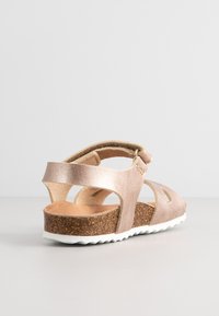 Roze gouden sandaal met een kurken zool, enkelbandje en uitsnijdingen. Het bovenwerk heeft een metallic afwerking en een gestreepte, gevoerde binnenkant.