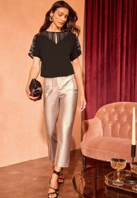 Top nero con dettagli metallici, scollatura a chiave; pantaloni slim-fit argento lucidi; pochette nera; décolleté nere con cinturini; sfondo di un divano rosa pallido.