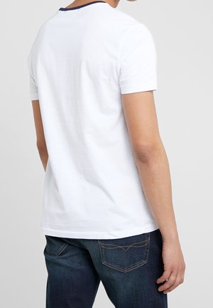 Mann trägt ein schlichtes weißes kurzärmeliges T-Shirt mit dunkelblauen Jeans, von hinten vor einem hellgrauen Hintergrund gezeigt.