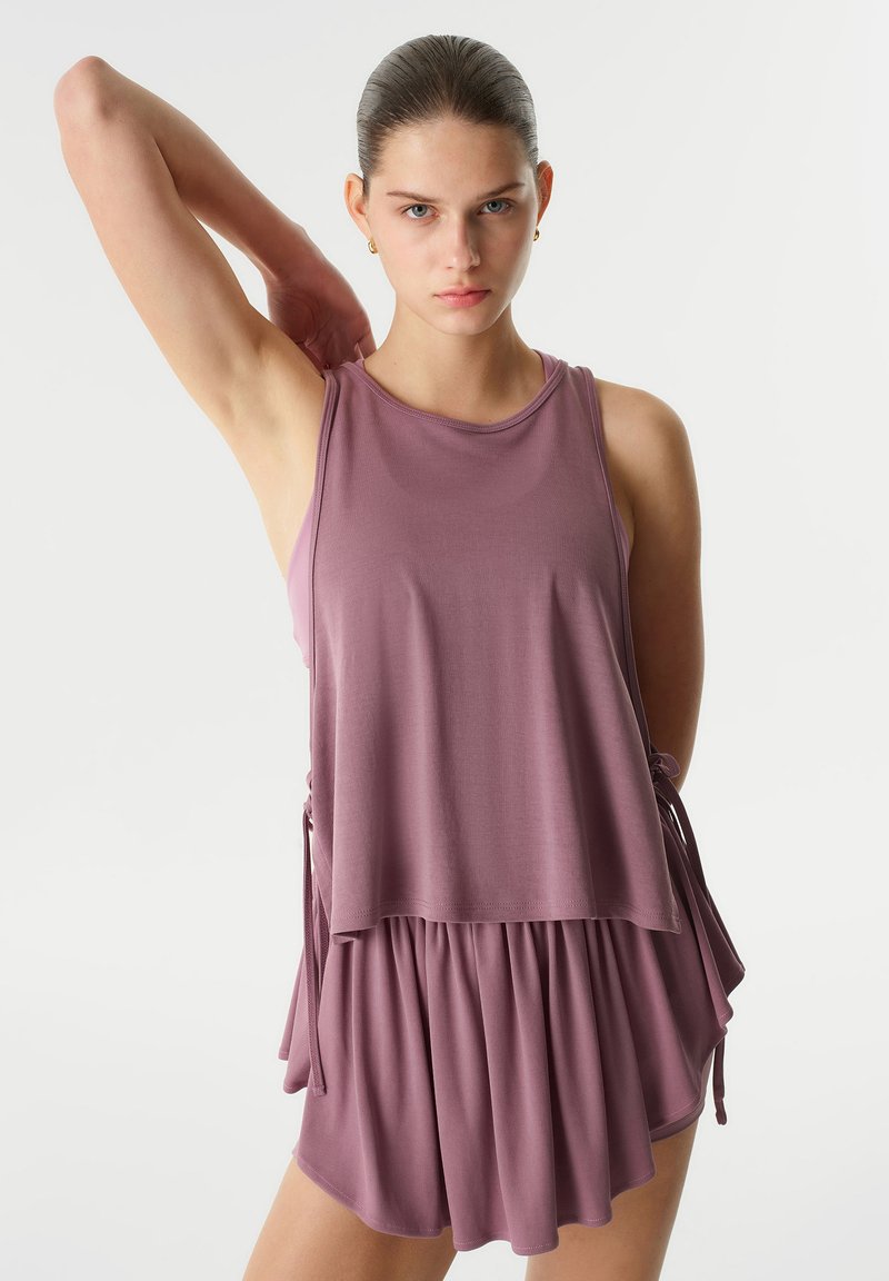 Conjunto atlético lila que incluye una blusa sin mangas con lazos laterales y una falda con capas, fabricado en una tela suave y ligera.