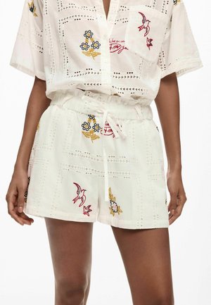 Mujer vistiendo pantalones cortos blancos y camisa con bordados de pájaros, flores y patrones de recortes tipo encaje, de pie con las manos relajadas a los lados.