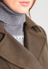 Abrigo verde oliva con gran cuello y solapa, superpuesto a un cuello de tortuga gris. Textura suave, costuras visibles y detalle estampado debajo.