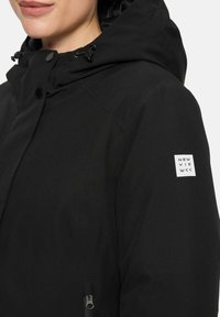 Vrouw in een zwarte capuchonjack met toggles en knopen, met een wit "NEW VIEW" logo op de bovenste mouw en een ritszak.