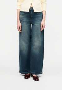 Άτομο που φοράει φαρδιά ξεβαμμένα μπλε τζιν, κρεμ πουλόβερ και σκούρα κόκκινα μπαλαρίνια από λουστρίνι, στέκεται μπροστά σε λευκό φόντο.