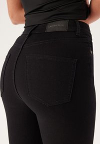 Czarne dżinsy o dopasowanym kroju, z tylnymi kieszeniami i subtelną metką marki. Gładka tekstura z mocnymi detalami szycia.
