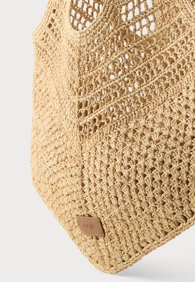 Sac cabas en paille tissée beige avec motif ajouré et poignée arrondie, comportant une petite étiquette en cuir cousue près du coin inférieur.