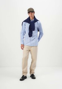 Camicia a righe azzurre e bianche, maglione blu navy appoggiato sulle spalle, pantaloni beige e scarpe nere; il cappello a quadri completa il look.