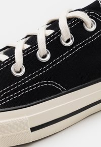 Primo piano di una sneaker nera in canvas con cuciture bianche, occhielli in metallo, lacci color crema e suola in gomma bianca con dettagli a striscia nera.