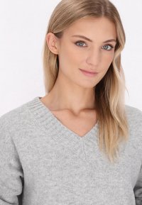 Jasnoszara sweter z dzianiny z dekoltem w kształcie litery V, z prążkowanym kołnierzem i miękką fakturą, zaprezentowany przez osobę z prostymi blond włosami.