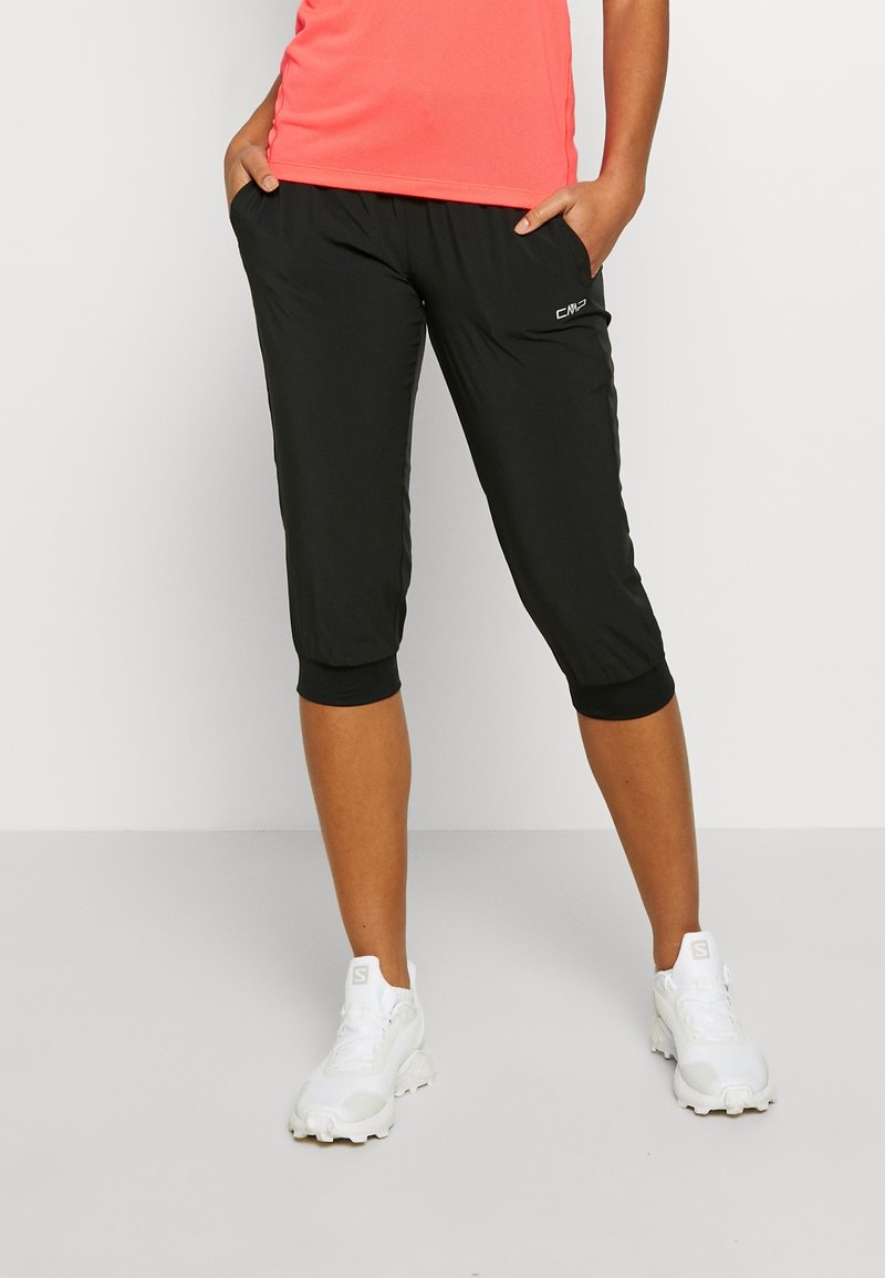 Personne portant un pantalon de sport court noir, un haut corail et des baskets blanches, debout, les mains dans les poches.