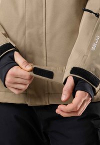 Beige Außensakko mit schwarzen Akzenten und Klettverschluss an den Bündchen, mit einer glatten Textur und verstärkten Nähdetails.