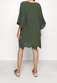 Olivgrünes Tunika-Kleid mit lockeren, ellbogenlangen Ärmeln und einem detaillierten Spitzenabschluss. Weicher Stoff mit entspannter Passform und rundem Halsausschnitt.