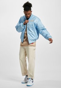 Veste bomber bleu clair avec des rayures verticales blanches, logo brodé, associée à un pantalon cargo beige et des baskets blanches avec des accents bleus.