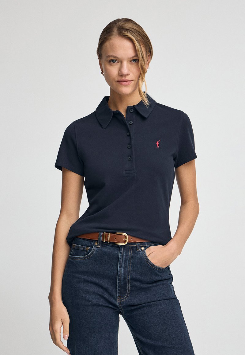 Dunkelblaues Polohemd mit klassischem Kragen, fünf Knöpfen und kleinem roten Logo. Getragen mit dunklen Jeans und einem braunen Gürtel.