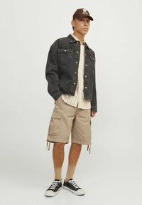 Sort denimjakke, beige cargo shorts, mønstret skjorte under, brun kasket, sorte sneakers med hvide såler, beige sokker. Afslappet outfit.