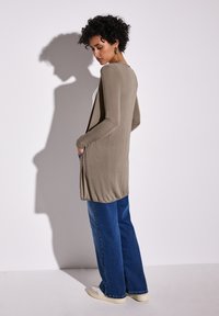 Cardigan lungo color marrone chiaro con maniche slim, due tasche laterali e orlo arrotondato, abbinato a jeans a gamba larga blu e scarpe da ginnastica bianche.