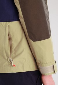 Kurtka z kapturem wykonana z khaki, granatowego materiału na tułowie i ciemnobrązową łatą na ramieniu. Posiada boczną kieszeń na zamek oraz regulację mankietów na rzep.