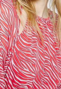 Blouse sans manches rouge avec un motif à rayures blanches ondulées, décolleté froncé et tissu léger. Vue rapprochée des détails du design.