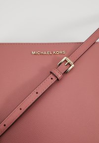 Różowa skórzana torebka o teksturowanym wykończeniu, z złotym logo "MICHAEL KORS" oraz regulowanym paskiem z metalową klamrą.