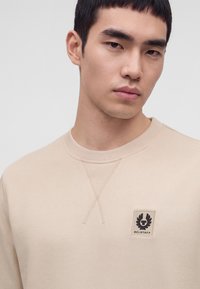 Млад мъж с къса тъмна коса, носещ бежова блуза с лого на Belstaff на гърдите, гледащ леко настрани.