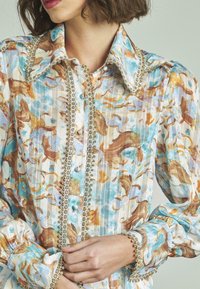 Camicia con motivo floreale nei colori azzurro chiaro, marrone e bianco. Caratterizzata da un tessuto testurizzato, un colletto ampio e un bordo decorativo con bottoni lungo la parte anteriore.