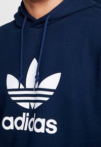 Person som bär en marinblå hoodie med vit Adidas Trefoil-logotyp och snoddar knutna i knoppar nära kragen.